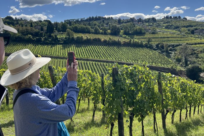 Hiker walking on a white gravel strade bianche road through Chianti Classico vineyards in Tuscany, with a medieval hilltop village in the background