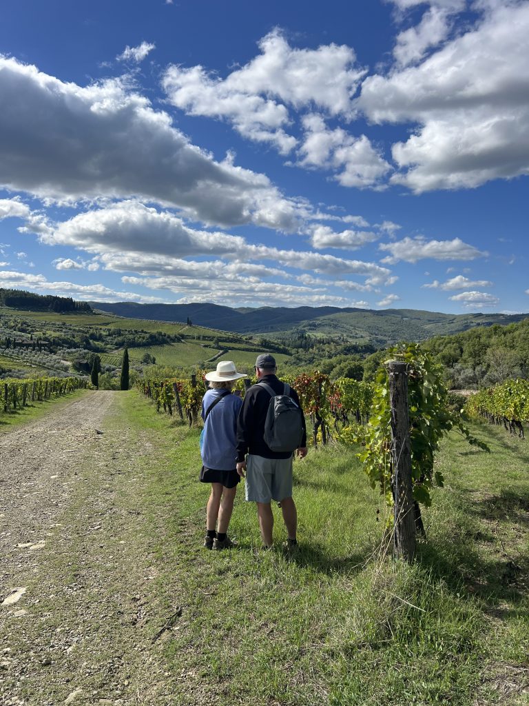 Hiker walking on a white gravel strade bianche road through Chianti Classico vineyards in Tuscany, with a medieval hilltop village in the background