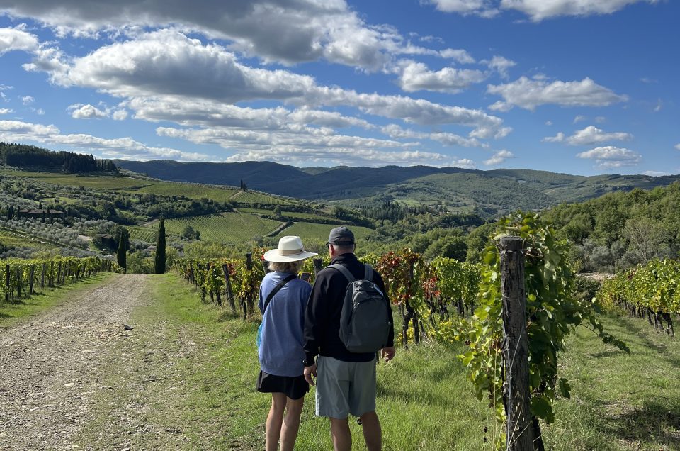 Hiker walking on a white gravel strade bianche road through Chianti Classico vineyards in Tuscany, with a medieval hilltop village in the background
